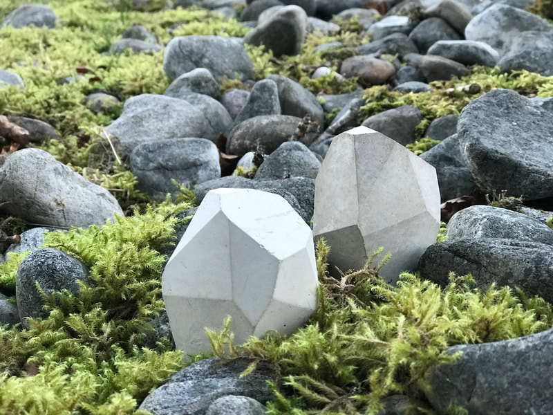 Photograph of two small stonecast geometric sculptures nestling on the shore amongst rocks and moss, made by Mara Marxt Lewis, entitled "Cast Stones", dated 2022, 
